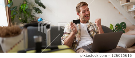 Portrait of cheerful guy buying flight tickets online, booking hotel by using laptop and credit card, sitting near suitcase in living room 126015616