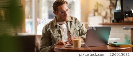 Portrait of handsome young guy in glasses, man studying, taking notes, looking at laptop screen during online lesson, attends a meeting and writing down information, sitting in cafe 126015681