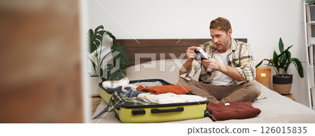 Portrait of young man looking at his digital camera screen, checking photo album on gadget, unpacking suitcase after holiday, travelling with luggage, sitting in hostel on a bed 126015835