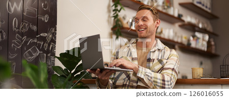 Image of young man in coffee shop, connects to video call, talking online, chats with friends via laptop and smiles Image of young man in coffee shop, connects to video call, talking online, chats with friends via laptop and smiles 126016055
