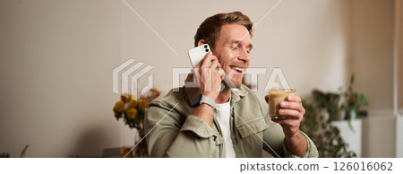 Portrait of young handsome man talking on the phone, drinks coffee and laughs. A guy sits in cafe having a nice, pleasant conversation over cup of cappuccino 126016062