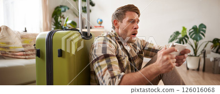 Man with shocked face, sitting on floor with suitcase, looking worried and tensed on mobile phone screen 126016068