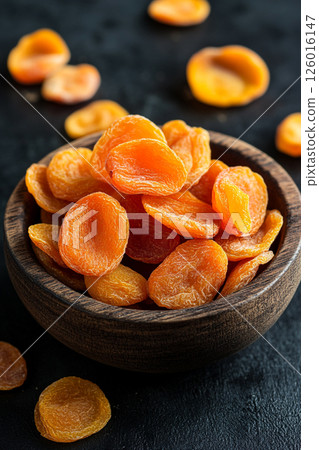 A wooden bowl brimming with dried apricots, some scattered around, their vibrant orange color contrasting beautifully with the dark, textured surface below. 126016147