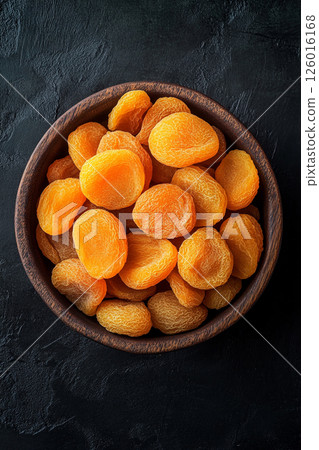 A wooden bowl brimming with dried apricots, some scattered around, their vibrant orange color contrasting beautifully with the dark, textured surface below. A wooden bowl brimming with dried apricots, some scattered around, their vibrant orange color contrasting beautifully with the dark, textured surface below. 126016168