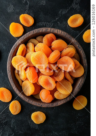 A wooden bowl brimming with dried apricots, some scattered around, their vibrant orange color contrasting beautifully with the dark, textured surface below. A wooden bowl brimming with dried apricots, some scattered around, their vibrant orange color contrasting beautifully with the dark, textured surface below. 126016169