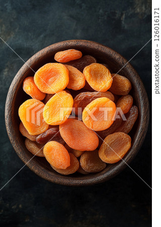 A wooden bowl brimming with dried apricots, some scattered around, their vibrant orange color contrasting beautifully with the dark, textured surface below. 126016171