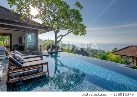 A luxurious seaside villa with an infinity pool overlooking the ocean, surrounded by lush greenery and featuring comfortable lounge chairs. 126016379