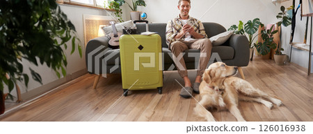 Portrait of happy, handsome young man with suitcase, sitting with his dog in living room, prepared to go on vacation, going on holiday Portrait of happy, handsome young man with suitcase, sitting with his dog in living room, prepared to go on vacation, going on holiday 126016938