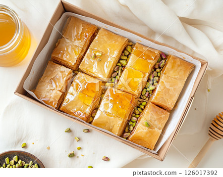 A tray of freshly baked baklava topped with chopped pistachios and honey, surrounded by bowls of honey and pistachios. The golden-brown pastry looks delicious and inviting. 126017392