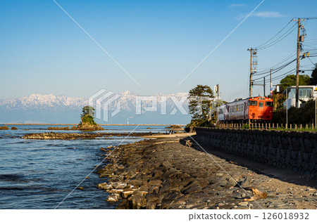 A JR Himi Line diesel train running along the coastline with the Onnaiwa Rocks of Amaharashi Coast and the snow-capped Tateyama mountain range in the background A JR Himi Line diesel train running along the coastline with the Onnaiwa Rocks of Amaharashi Coast and the snow-capped Tateyama mountain range in the background 126018932