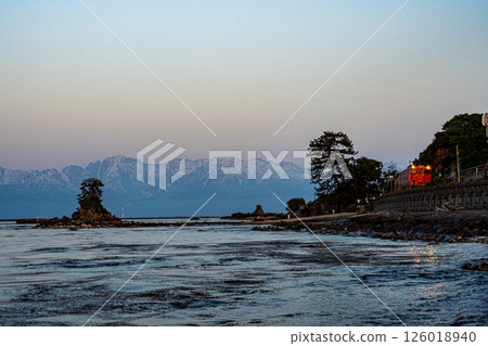 At dusk on Amaharashi Beach, a JR Himi Line diesel train runs along the coastline with the Onnaiwa Rock floating in Toyama Bay and the snow-capped Tateyama mountain range in the background. At dusk on Amaharashi Beach, a JR Himi Line diesel train runs along the coastline with the Onnaiwa Rock floating in Toyama Bay and the snow-capped Tateyama mountain range in the background. 126018940