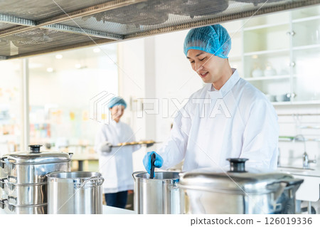 A male chef wearing a sanitary white coat cooking in a large pot in the kitchen, school lunch center, cafeteria A male chef wearing a sanitary white coat cooking in a large pot in the kitchen, school lunch center, cafeteria 126019336