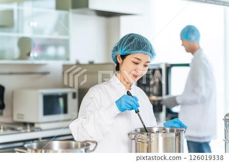 A female chef wearing a sanitary white coat cooking in a large pot in the kitchen, school lunch center, cafeteria A female chef wearing a sanitary white coat cooking in a large pot in the kitchen, school lunch center, cafeteria 126019338