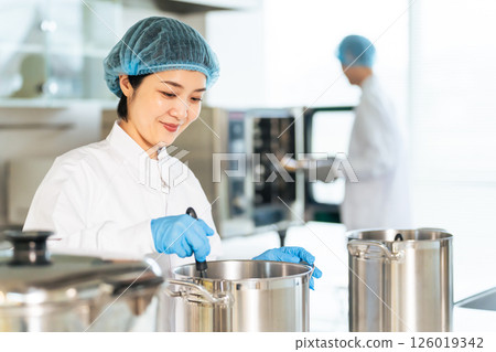 A female chef wearing a sanitary white coat cooking in a large pot in the kitchen, school lunch center, cafeteria A female chef wearing a sanitary white coat cooking in a large pot in the kitchen, school lunch center, cafeteria 126019342
