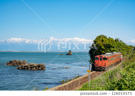 A JR Himi Line diesel train running along Amaharashi Coast with Otokoiwa Rock floating in Toyama Bay and the snow-capped Tateyama mountain range in the background A JR Himi Line diesel train running along Amaharashi Coast with Otokoiwa Rock floating in Toyama Bay and the snow-capped Tateyama mountain range in the background 126019373