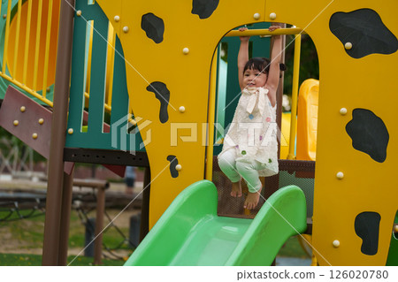 happy toddler girl hanging the yellow bar by her hand at outdoor playground in park 126020780