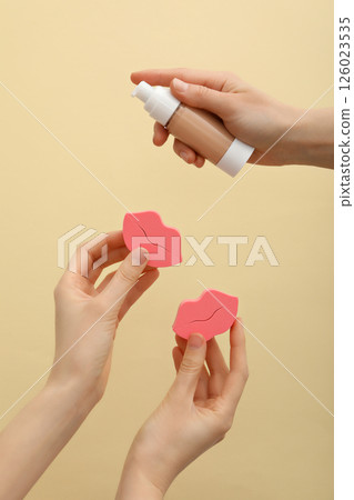 Bottle of foundation and cosmetic sponges in female hands on beige background 126023535