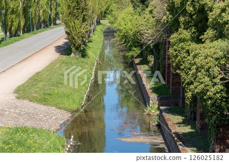 A deep moat at the foot of the massive stone walls of an ancient castle. Medieval architecture, a defensive structure surrounded by greenery and traces of time. A deep moat at the foot of the massive stone walls of an ancient castle. Medieval architecture, a defensive structure surrounded by greenery and traces of time. 126025182