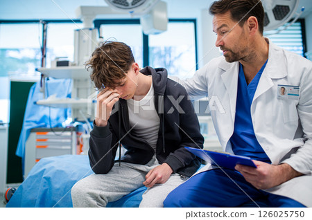 Depressed teenage patient sitting on hospital bed, talking with caring doctor. 126025750