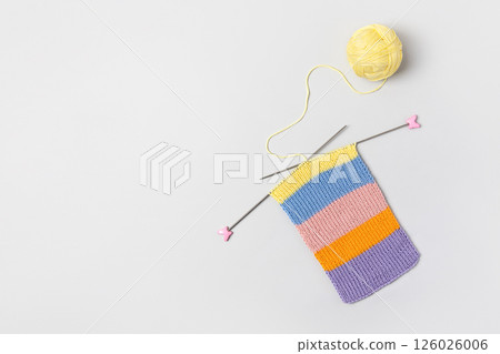 Striped piece of knitted fabric for future clothing with knitting needles and yellow ball of thread on gray background. Homemade needlework and knitting of warm, clothes and toys. Concept creativity Striped piece of knitted fabric for future clothing with knitting needles and yellow ball of thread on gray background. Homemade needlework and knitting of warm, clothes and toys. Concept creativity 126026006