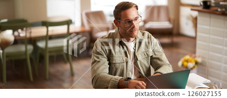 Portrait of young concentrated man in glasses, working from coffee shop, studying, looking thoughtful while completing task on laptop, making notes in notebook Portrait of young concentrated man in glasses, working from coffee shop, studying, looking thoughtful while completing task on laptop, making notes in notebook 126027155