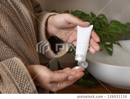 Woman in brown bathrobe applying cream on fingers and holding cosmetic tube in other hand, mockup Woman in brown bathrobe applying cream on fingers and holding cosmetic tube in other hand, mockup 126031138