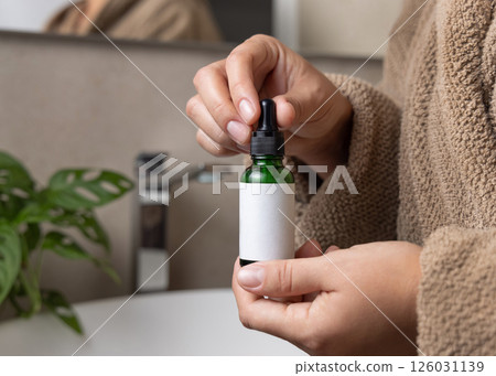 Hands in brown bathrobe holding green dropper bottle with label closeup, cosmetic mockup 126031139