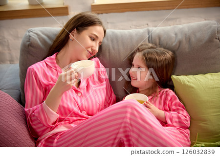 Mother and daughter in pink pajamas, sitting on couch in living room at home, drinking tea and sharing warm moment together. 126032839