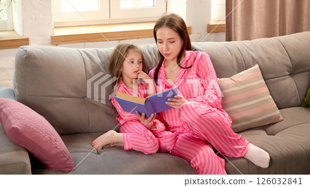 Mother and daughter enjoying quiet moment together at home, sitting on couch in pink pajamas and reading book. 126032841