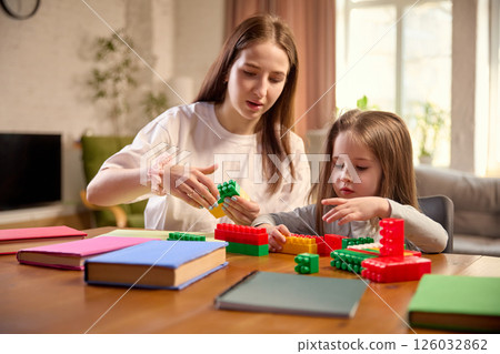 Mother and daughter engaged in shared building block activity, sitting at table in living room at home Mother and daughter engaged in shared building block activity, sitting at table in living room at home 126032862