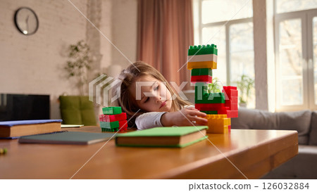 Child girl sitting at desk at home in living room, playing with building blocks and notebooks. 126032884