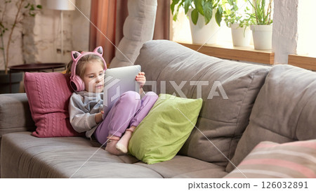 Young child comfortably lying on couch with headphones on and tablet in hand, focused on screen. Young child comfortably lying on couch with headphones on and tablet in hand, focused on screen. 126032891