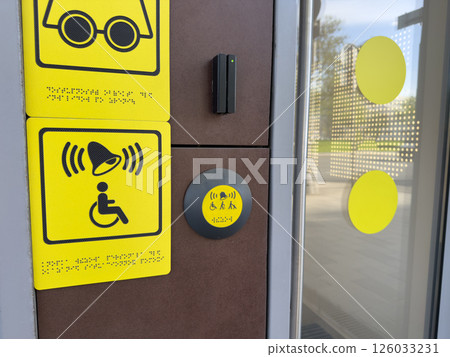 Accessibility call system with yellow tactile and audio symbols, including Braille text and assistance button near glass door, designed to support people with disabilities in modern infrastructure 126033231