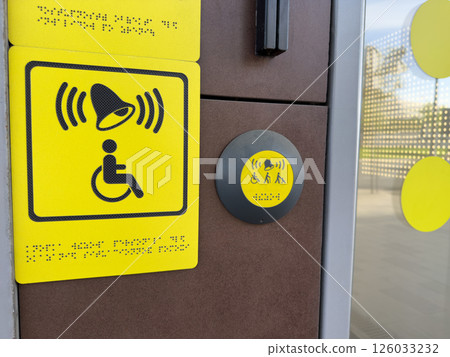 Yellow tactile signage and assistance button with bell and disability symbols including Braille, providing accessible user interface near modern entrance for inclusive and barrier free access 126033232