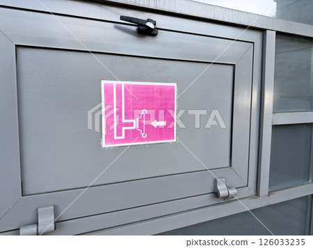 Close up of gray metal cabinet with pink fire hose reel sign showing schematic icon and directional arrow, part of exterior fire safety system installed on public or commercial building facade 126033235