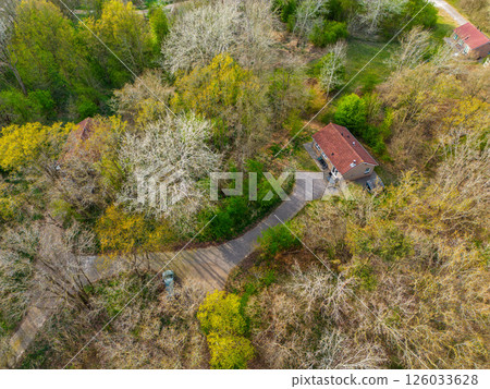 Top-down aerial view of a small house with a red-tiled roof, surrounded by a forest with trees in early spring colors, and a narrow driveway leading to a parked car. Top-down aerial view of a small house with a red-tiled roof, surrounded by a forest with trees in early spring colors, and a narrow driveway leading to a parked car. 126033628