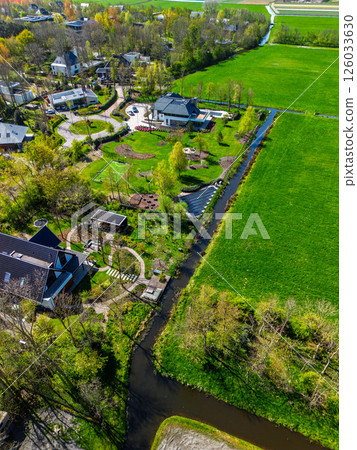 Aerial view of a modern countryside neighborhood with green lawns, small canals, landscaped gardens, and residential houses surrounded by fields and patches of forest. 126033630