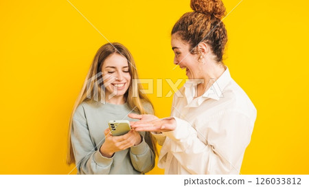Two young women sharing funny content on a smartphone on yellow background 126033812