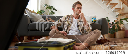 Portrait of stressed young man packing his suitcase, deciding what clothes bring on vacation, thinking while looking at luggage Portrait of stressed young man packing his suitcase, deciding what clothes bring on vacation, thinking while looking at luggage 126034019