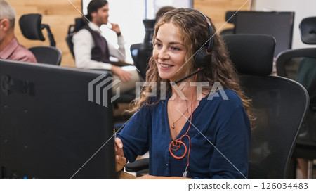 Happy businesswoman looking at computer as she wears headset in coworking office Happy businesswoman looking at computer as she wears headset in coworking office 126034483