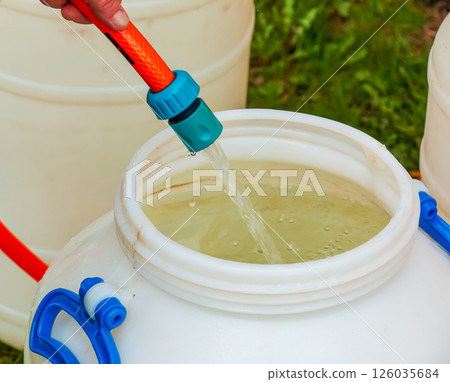 A woman's hand fills a plastic barrel with water from a hose. The concept of the drinking water crisis in the world. A woman's hand fills a plastic barrel with water from a hose. The concept of the drinking water crisis in the world. 126035684