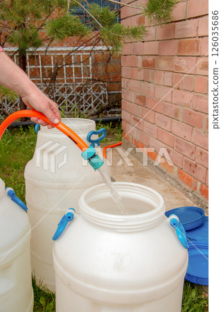 A woman's hand fills a plastic barrel with water from a hose. The concept of the drinking water crisis in the world. A woman's hand fills a plastic barrel with water from a hose. The concept of the drinking water crisis in the world. 126035686