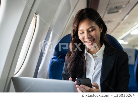 Business travel woman smiling using phone and laptop in airplane Business travel woman smiling using phone and laptop in airplane 126036264