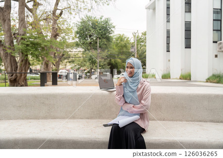 Muslim businesswoman sitting outdoors with a laptop and notebook, smiling. 126036265
