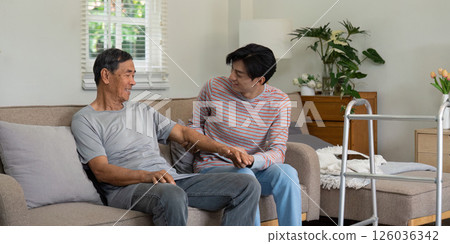 Joyful son sharing a moment with elderly father on the couch, highlighting family bonds and support in a cozy living room. 126036342