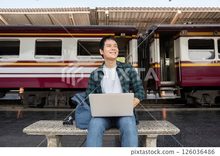 Travel and Technology. A young traveler working on his laptop at a train station, excited for his next adventure. Travel and Technology. A young traveler working on his laptop at a train station, excited for his next adventure. 126036486