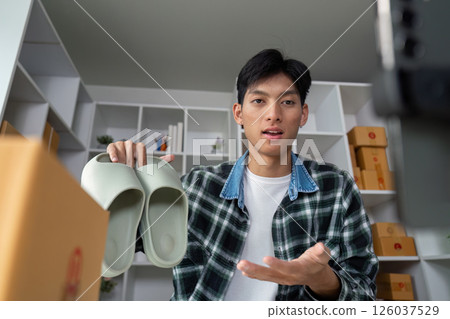 Young entrepreneur showcasing footwear during online presentation, holding shoes in modern workspace with product boxes. 126037529