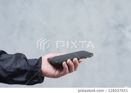 A man in a black jacket holding a smartphone against a gray concrete wall. Modern technology and the use of mobile devices. 126037831