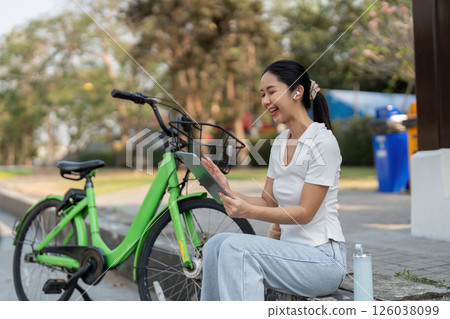 Nature and Digital Interaction. A woman connects with her tablet while taking a break beside her bicycle in a park. Nature and Digital Interaction. A woman connects with her tablet while taking a break beside her bicycle in a park. 126038099