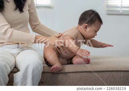 Mother helping baby sit up on a couch, fostering early development and bonding. Mother helping baby sit up on a couch, fostering early development and bonding. 126038219